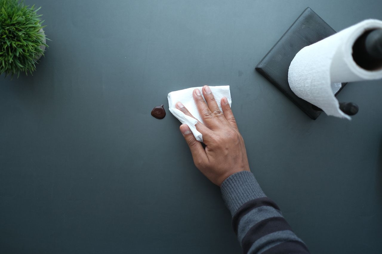 Une main nettoie une surface sombre avec un mouchoir, près d'un rouleau de papier et d'une plante.